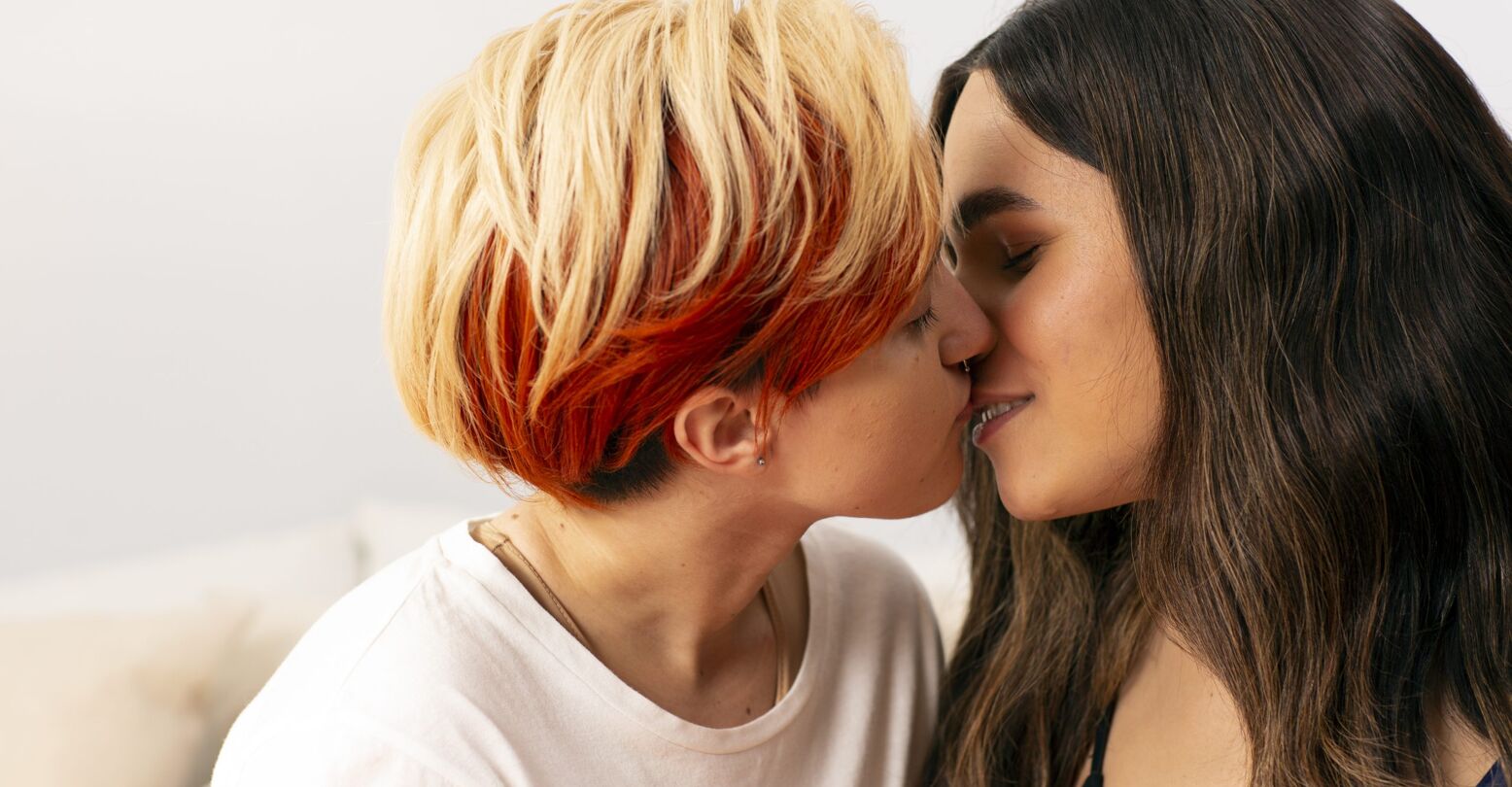 A transmasculine gender-nonconforming person and transfeminine non-binary person kissing