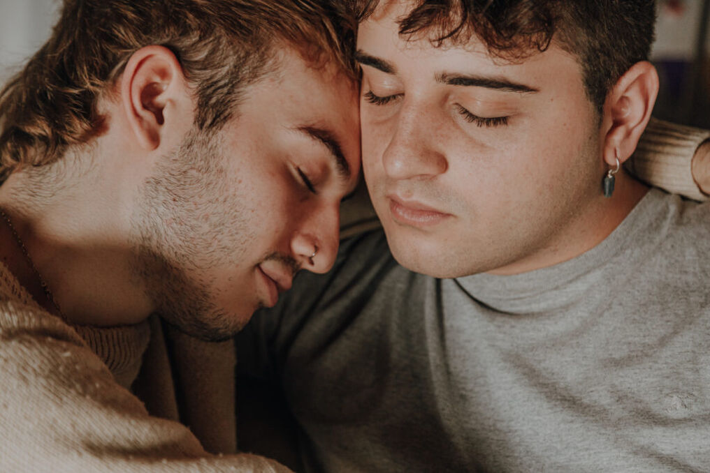 two male-presenting people cuddle closely together with their eyes closed
